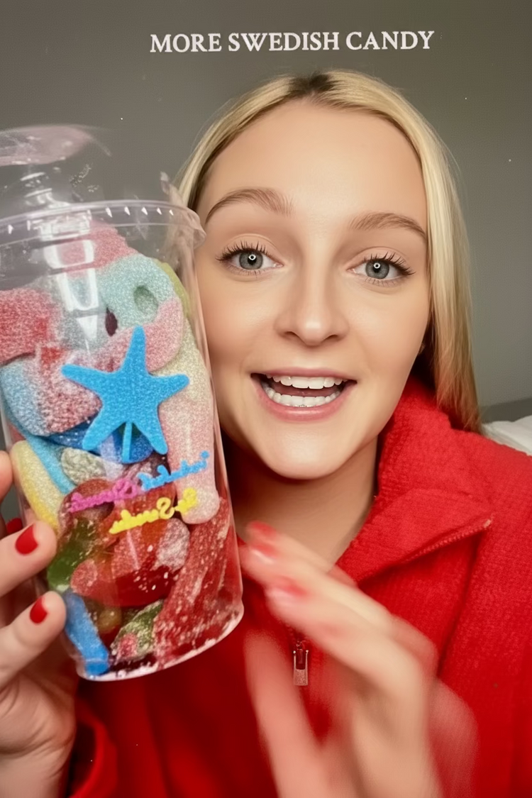 A smiling blonde woman in a red fleece jacket holds up a clear plastic cup filled with colorful Swedish gummy candies, including a blue starfish-shaped candy and rainbow gummies. The text 'MORE SWEDISH CANDY' is written at the top against a gray background.