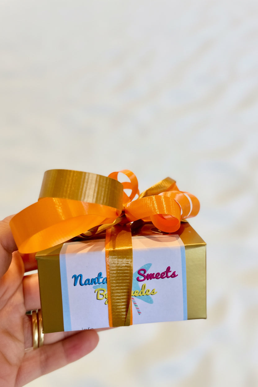A box of Petit Pralines chocolate with an orange ribbon, presented on a glass shelf with various other chocolate items in the background.
