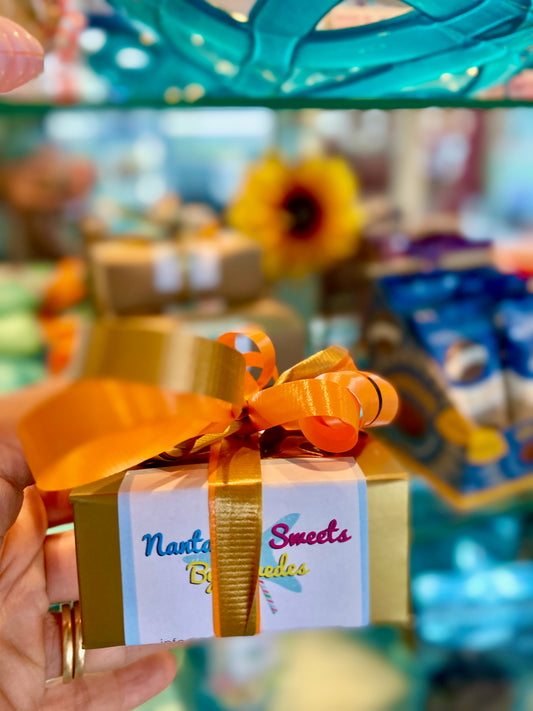 A box of Petit Pralines chocolate with an orange ribbon, presented on a glass shelf with various other chocolate items in the background.