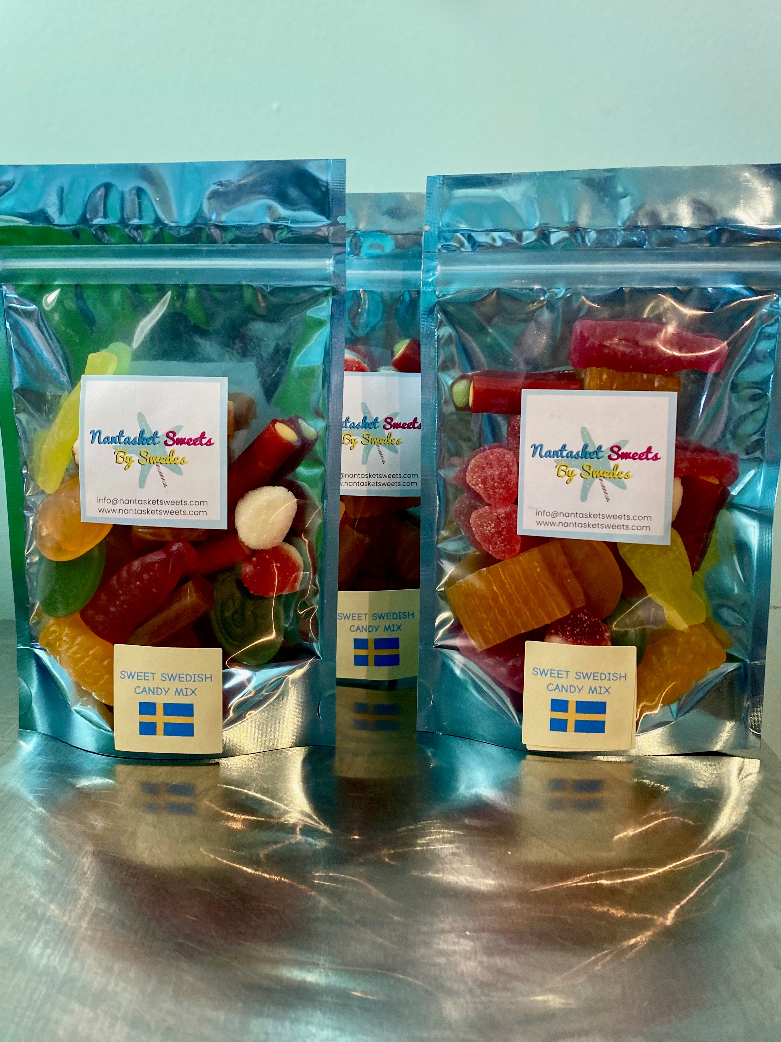 Three packages of 'Sweet Swedish' gummy candies on a reflective surface with a light blue background.
