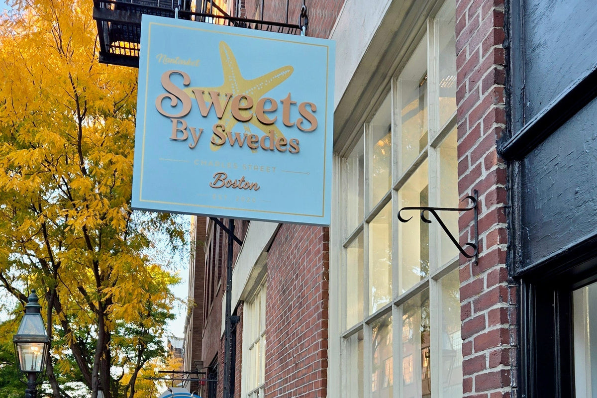 A street-side view of a brick storefront with a hanging sign reading 'Sweets by Swedes' .' A sidewalk runs alongside the building, with autumn trees showing yellow leaves and a pedestrian in a red jacket walking below.