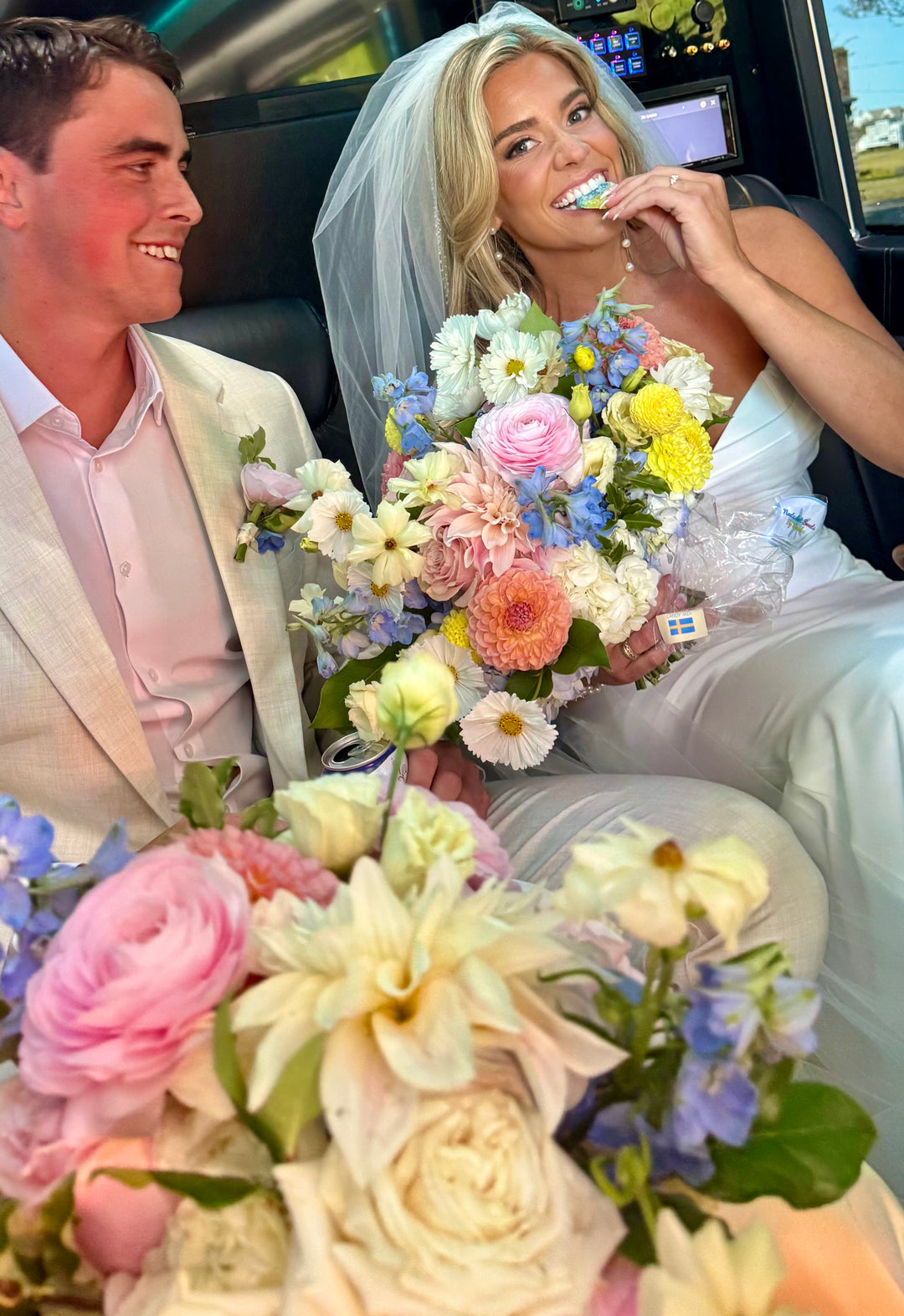 Woman in a wedding dress holding a bouquet of flowers, sitting next to a man in a suit inside a car.